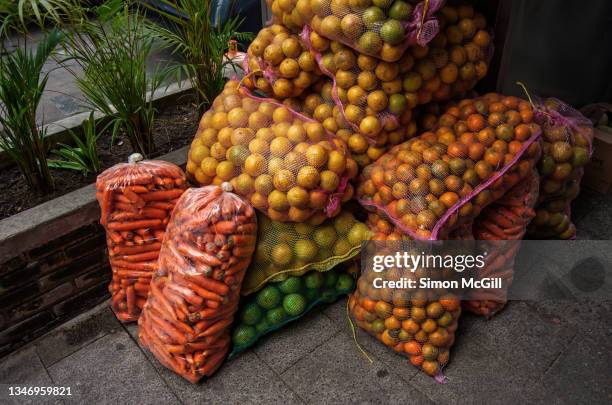 mesh bags of oranges and plastic bags of carrots sitting in a heap on the sidewalk next to a fruit juice stall - tela ruvida foto e immagini stock