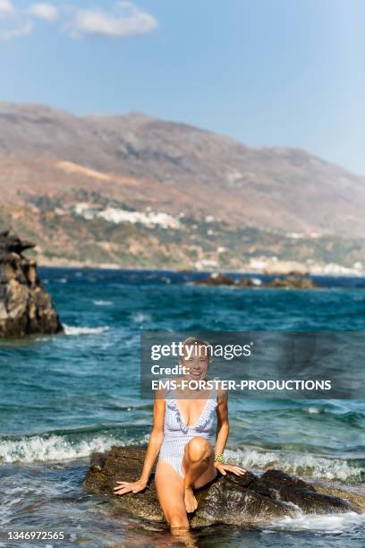 smiling, mature woman is sitting and relaxing on a rock in the sea - crete woman stock pictures, royalty-free photos & images