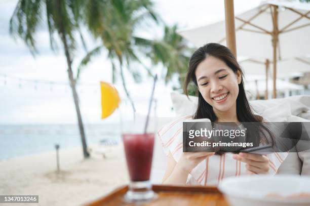 asiatische frau synchronisieren einer kreditkarte mit einem technischen gerät wie einem smartphone oder tablet tippen einer kreditkarte auf eine lesetechnologie als kreditkartenersatz am tropischen strand in südostasien - south east asia stock-fotos und bilder