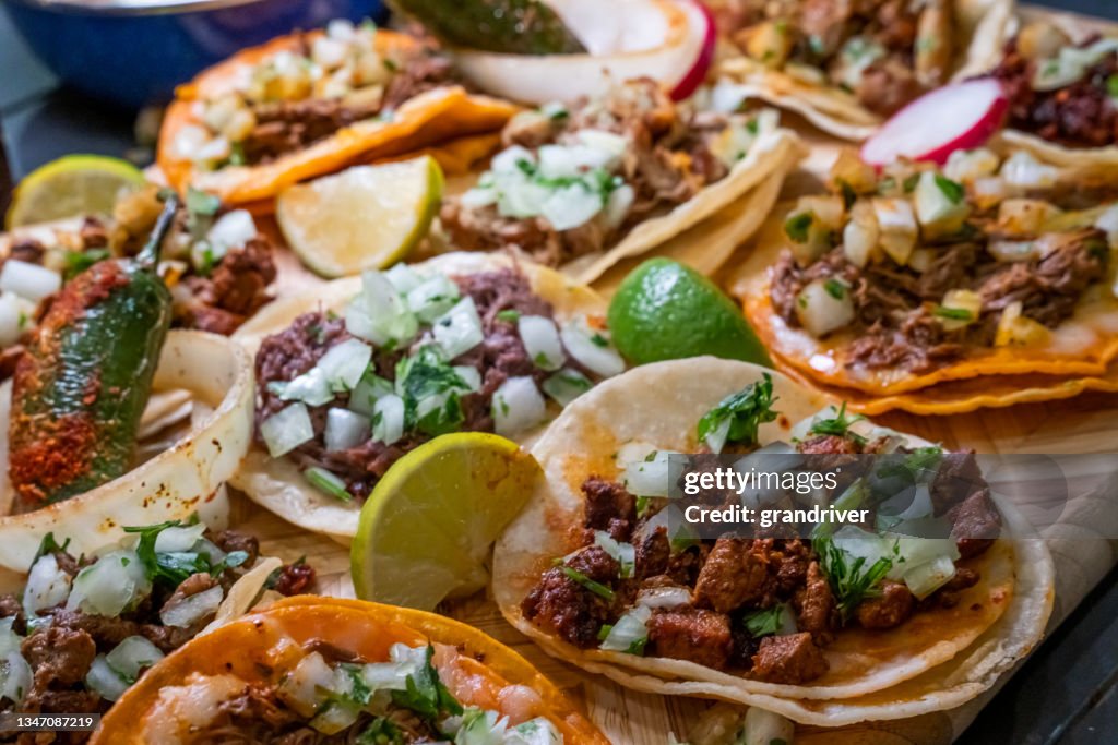 Auswahl an köstlichen authentischen Tacos, Birria, Carne Asada, Adobada, Cabeza und Chicharone, arrangiert mit Limettenscheiben, Zwiebeln und geröstetem Chilischoten