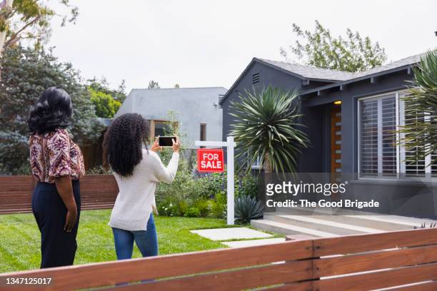 young woman photographing house through mobile phone with mother - house with for sale sign stock-fotos und bilder