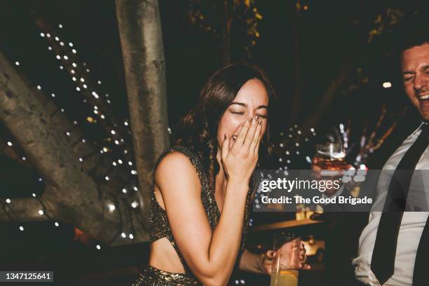 cheerful woman covering mouth with hand while standing by male friend at party - cóctel fiesta fotografías e imágenes de stock