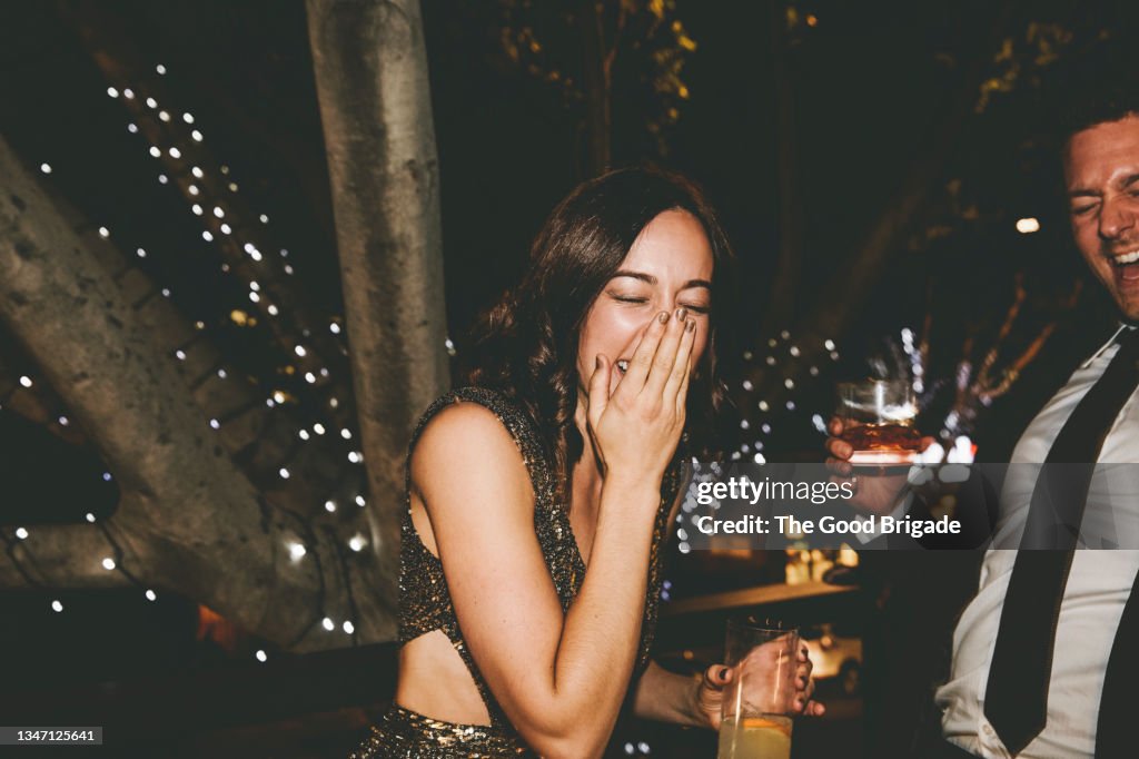 Cheerful woman covering mouth with hand while standing by male friend at party