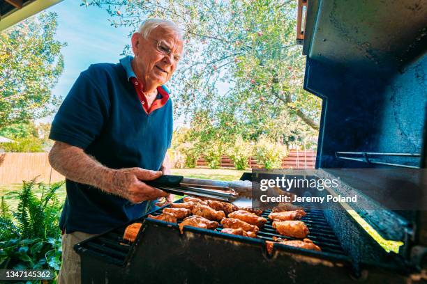 cheerful senior caucasian man standing outdoors on the back patio checking the doneness of spicy, delicious buffalo chicken wings on the grill for a summer afternoon lunch or dinner party - bbq chicken wings stock pictures, royalty-free photos & images