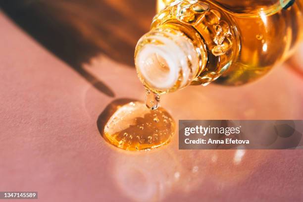 drop of bright yellow essential oil with air bubbles is dripping from glass bottle on pastel pink background. play of shadow and light creates an abstract pattern. trendy colors of the year 2021. extreme close-up - argan oil stock-fotos und bilder