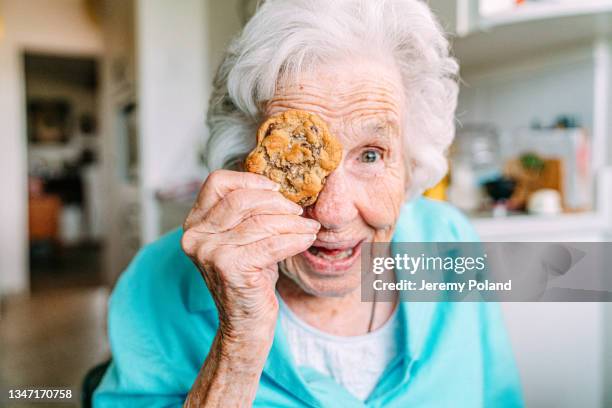 süßes porträt einer fröhlichen, scharfen 100-jährigen seniorin frau, die einen süßen keks vor ihrem auge hält und ein gesicht in die kamera lächeln lässt - 100 jahre alter mensch stock-fotos und bilder