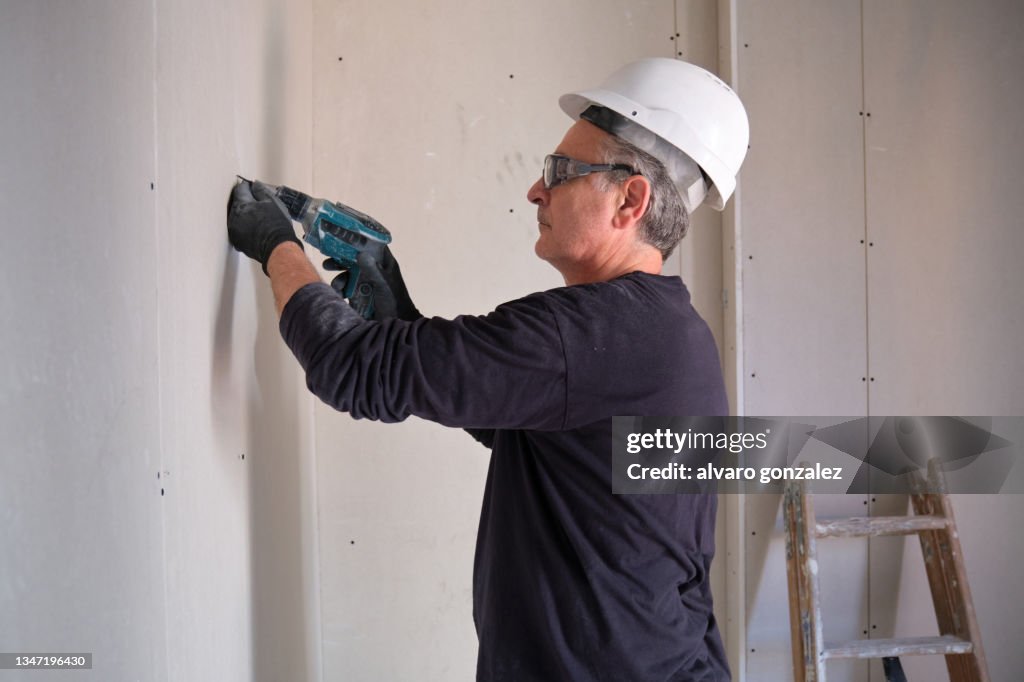 Builder screws a plasterboard wall while working in the interior of a construction site.