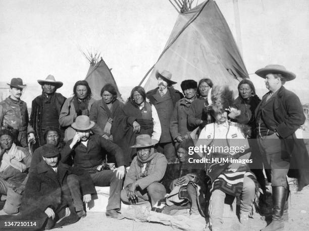 Group of White Americans and Lakota men standing and sitting in two rows in front of tipis..