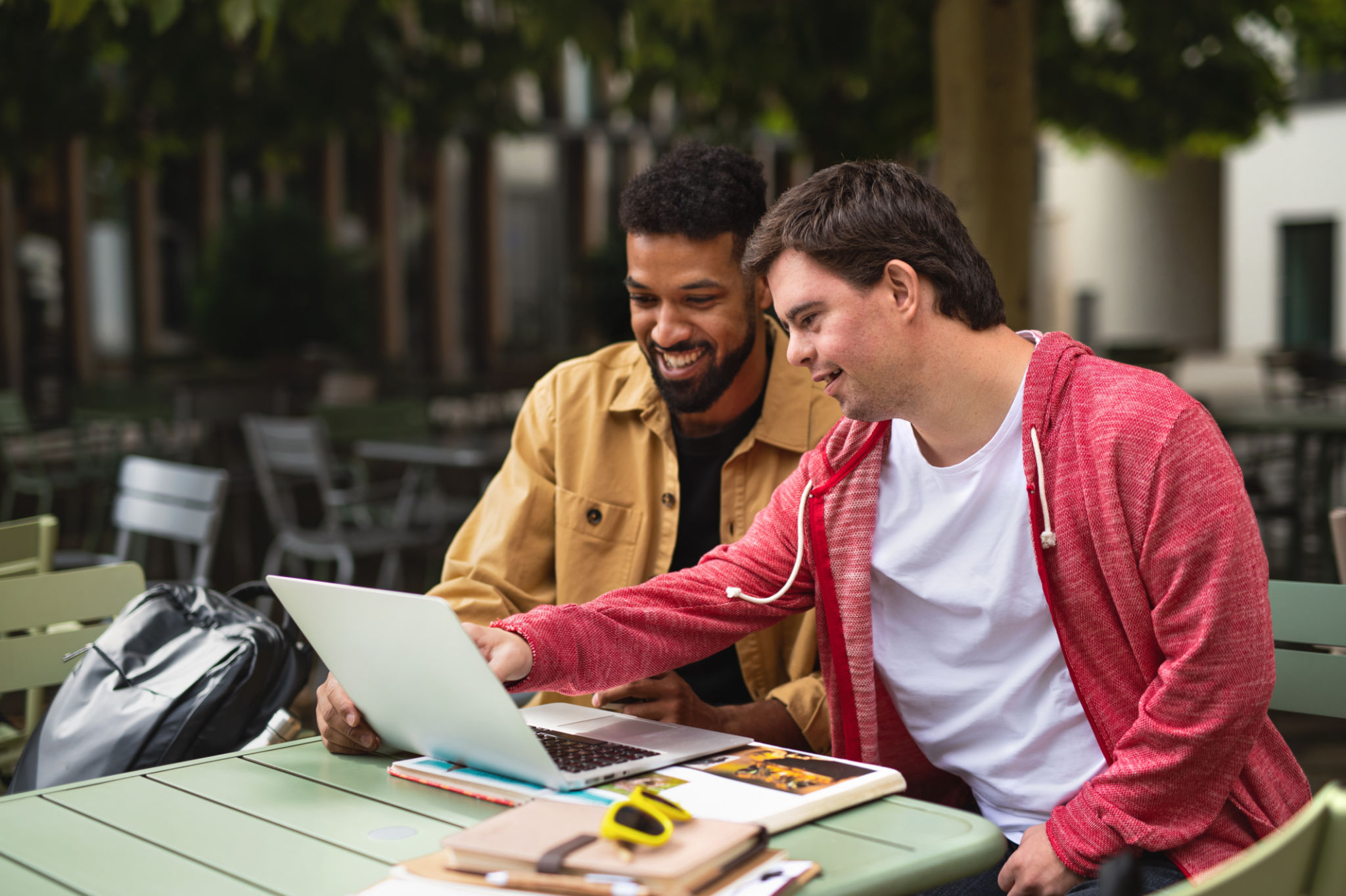 Young man with Down syndrome with his mentoring friend sitting outdoors in cafe using laptop. Young man with Down syndrome with his mentoring friend sitting outdoors in cafe using laptop.
