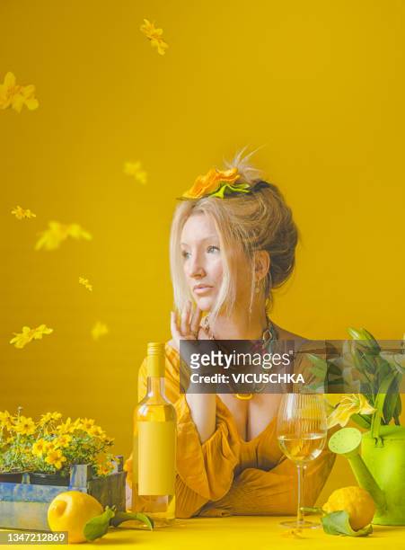woman in yellow dress with headdress, colorful necklace at table with potted flowers, wine bottle with empty label mock up, wine glass, pears and watering can on yellow wall background - flower necklace stock pictures, royalty-free photos & images