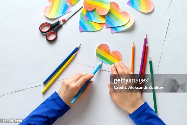 a girl or woman paints a heart cut out of paper in the color of the lgbt flag. next to it are bright colored pencils, against the background of a wooden table. valentine in the color of the rainbow. the concept of lgbt and human rights, valentine's day. - child cutting card stock-fotos und bilder