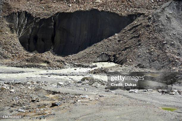 The snout of a glacier carrying morraine, with large blocks of ice broken off and a meltwater river flowing out.