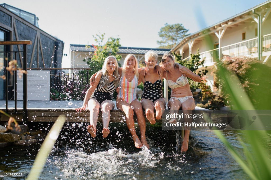 Laughing senior female swimmers sitting on jetty preparing for early morning swim in hotel resort.