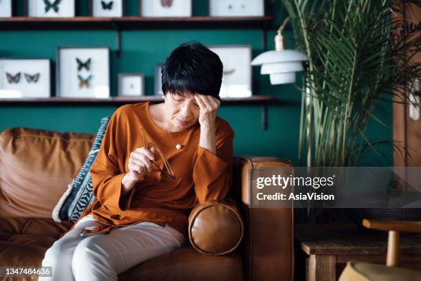 una donna asiatica anziana sconvolta che si sente male, soffre di mal di testa mentre è seduta sul divano nel soggiorno di casa - menopausa foto e immagini stock