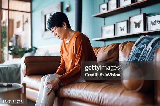 A distraught senior Asian woman feeling unwell, suffering from pain in leg while sitting on sofa in the living room at home A distraught senior Asian woman feeling unwell, suffering from pain in leg while sitting on sofa in the living room at home