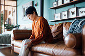 A distraught senior Asian woman feeling unwell, suffering from pain in leg while sitting on sofa in the living room at home