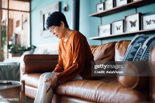 a distraught senior asian woman feeling unwell, suffering from pain in leg while sitting on sofa in the living room at home - exaustão imagens e fotografias de stock