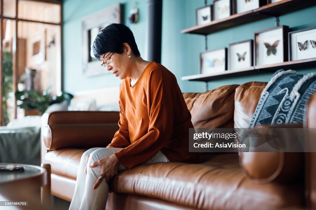 A distraught senior Asian woman feeling unwell, suffering from pain in leg while sitting on sofa in the living room at home