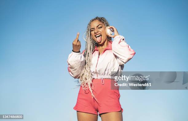 woman showing obscene gesture while listening music through wireless headphones - v sign stock pictures, royalty-free photos & images