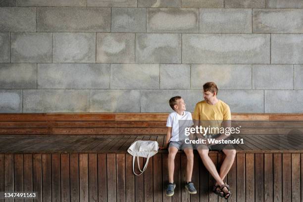 smiling son and father looking at each other while sitting on bench - família de duas gerações imagens e fotografias de stock