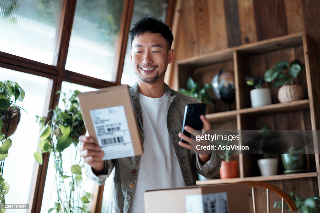 Lächelnder junger Asiatischer, der elektronisches Banking auf seinem Smartphone überprüft, als er gelieferte Pakete von Online-Einkäufen zu Hause erhielt. Online-Shopping. Online-Banking. Sicher online einkaufen und bezahlen