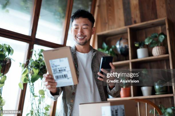 smiling young asian man checking electronic banking on his smartphone as he received delivered packages from online purchases at home. online shopping. online banking. shopping and paying safely online - e commerce payment stockfoto's en -beelden