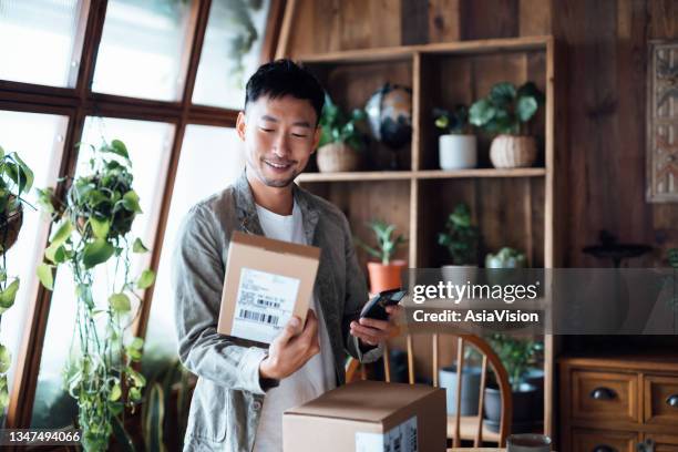 smiling young asian man with smartphone, receiving delivered packages from online purchases at home, can't wait to unbox the purchases. online shopping, enjoyable customer shopping experience - online shop stockfoto's en -beelden