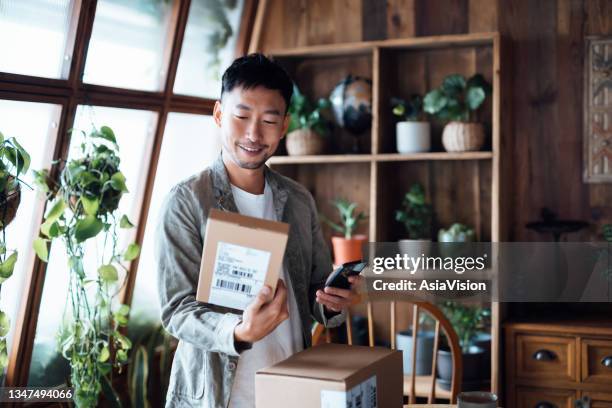 smiling young asian man with smartphone, receiving delivered packages from online purchases at home, can't wait to unbox the purchases. online shopping, enjoyable customer shopping experience - compras em casa imagens e fotografias de stock