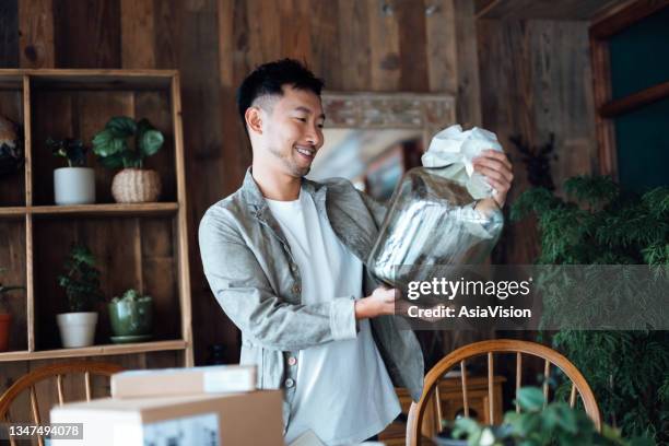 smiling young asian man looking happy as he unboxing the delivered package from online purchases at home, take out a glass vase. online shopping, enjoyable customer shopping experience - chinese young adults shopping stock pictures, royalty-free photos & images