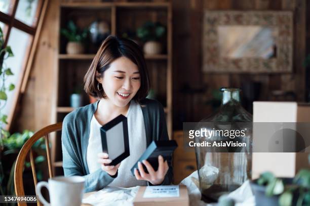 mulher asiática sorridente parece feliz enquanto desboxing os pacotes entregues de compras online em casa. compras online, experiência de compra de clientes agradável - unboxing - fotografias e filmes do acervo