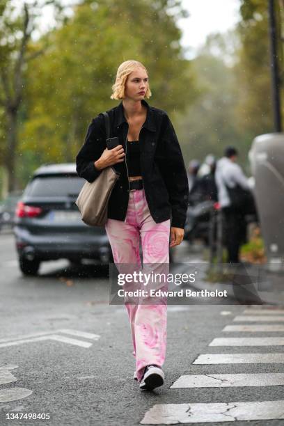 Model wears a black V-neck cropped tank-top, a black oversized zipper coat, a black shiny leather belt, white and pink tie and dye flared pants, a...