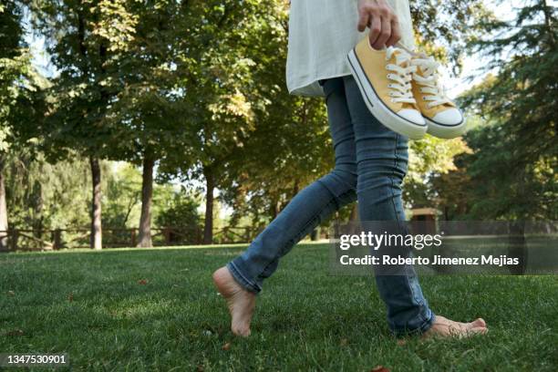 woman walking barefoot on grass, holding her sneakers. lower section - barefoot foto e immagini stock