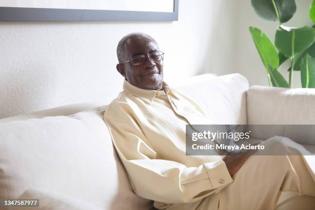 portrait of an african american senior man with grey hair and glasses, sits in the sofa in a brightly lit living room wearing beige colored top and pants. - beige pants - fotografias e filmes do acervo