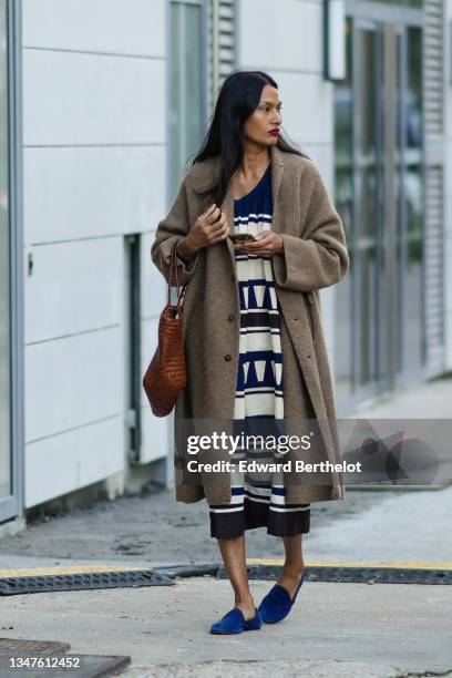 Guest wears a brown wool long oversized coat, a navy blue and white striped pattern long flowing midi dress, a brown shiny braided leather handbag,...