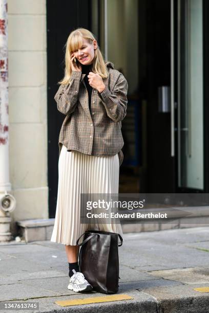 Guest wears a black turtleneck pullover, a brown and orange small checkered oversized shirt, a white latte pleated / accordion midi skirt, black...