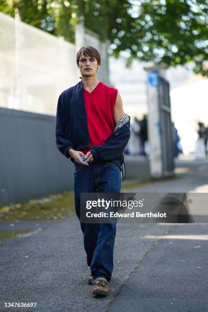 Guest wears a red wool sleeveless pullover, a navy blue wool coat with checkered pattern interior, navy blue denim jeans large pants, brown faded...