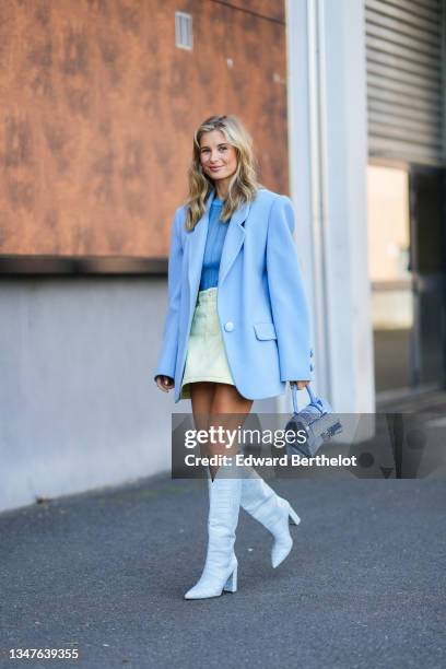 Xenia Adonts wears large silver earrings, a blue ribbed t-shirt, a pale yellow denim short skirt, a pale / baby blue oversized blazer jacket, a blue...