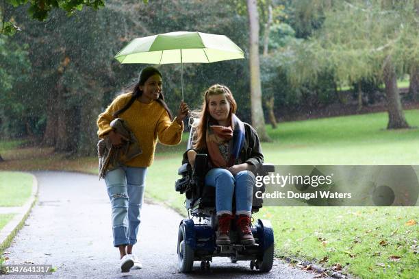 woman in wheelchair and friend with umbrella walking through park in rain. - mobility disability stock pictures, royalty-free photos & images