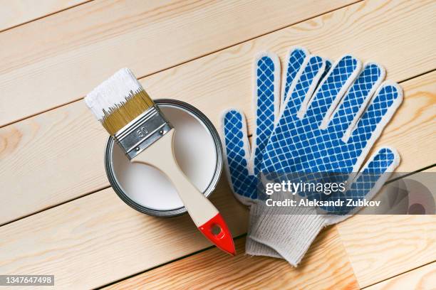 brush in white paint on a can of paint, on a wooden background. new protective gloves are next to it. painting and repair work. the concept of the construction industry, professional and home repair. - lackiert stock-fotos und bilder