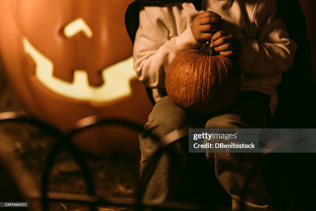 Child sitting with a pumpkin at night on a Halloween party with a Jack-o-Lantern lit in the background. Concept of fear and celebration of Halloween and carnival
