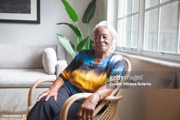 portrait of an african american senior woman, with white hair, happily sits in a brightly lit living room wearing a colorful blouse. - woman home sit foto e immagini stock