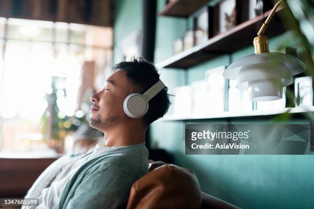 young asian man with eyes closed, enjoying music over headphones while relaxing on the sofa at home - tevreden stockfoto's en -beelden