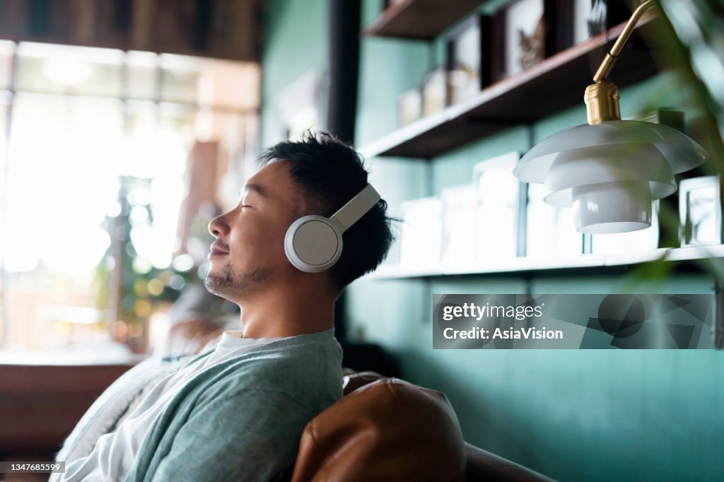 Young Asian man with eyes closed, enjoying music over headphones while relaxing on the sofa at home