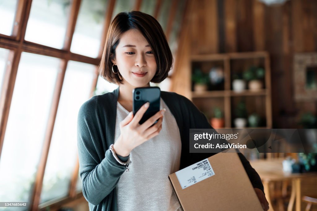 Smiling young Asian woman shopping online with smartphone on hand, receiving a delivered parcel by home delivery service. Online shopping, online banking. Enjoyable customer shopping experience