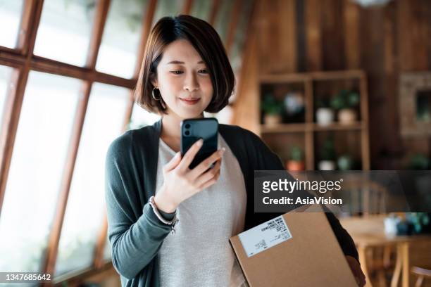 smiling young asian woman shopping online with smartphone on hand, receiving a delivered parcel by home delivery service. online shopping, online banking. enjoyable customer shopping experience - debit cards credit cards accepted stock pictures, royalty-free photos & images