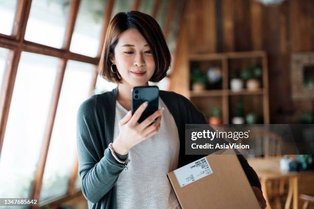 smiling young asian woman shopping online with smartphone on hand, receiving a delivered parcel by home delivery service. online shopping, online banking. enjoyable customer shopping experience - returning bildbanksfoton och bilder