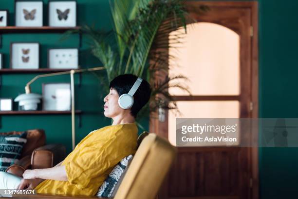 mujer asiática mayor con los ojos cerrados, disfrutando de la música con auriculares mientras se relaja en el sillón de casa - headphones asian fotografías e imágenes de stock