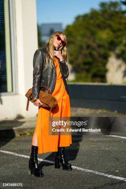 Guest wears vintage red sunglasses, a large chain necklace, gold chain pendant necklace, an orange shiny flowing midi dress, a black shiny leather...