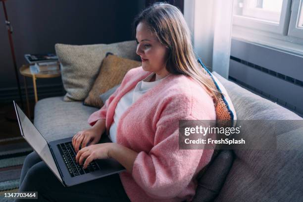 beautiful overweight woman sitting on the sofa in her living room and browsing the web on a laptop - body confidence stock pictures, royalty-free photos & images