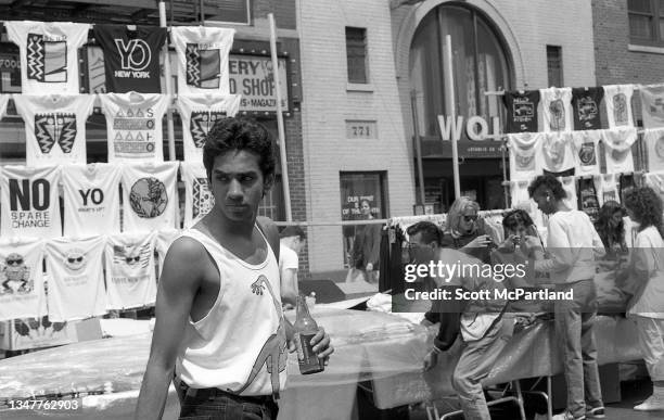 Man holds a bottled drink as he walks on 9th Avenue while, behind him, other pedestrians browse through t-shirts at vendor's stalls in Hell's Kitchen...